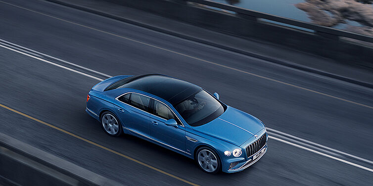 Bentley Lima New Bentley Flying Spur Azure sedan in Light windsor blue paint by Mulliner, with 22 inch Azure wheels seen from overhead driving dynamically on a country road